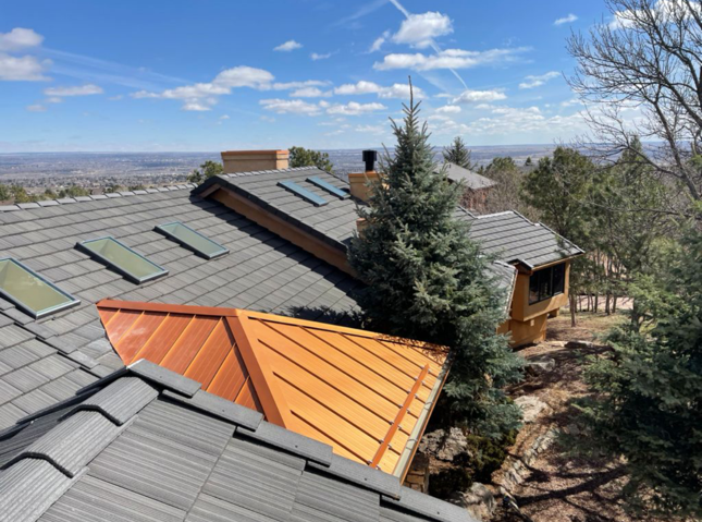 House with gray roof tiles, copper roof accent, skylights, and evergreen trees on a sunny day with a distant city view.