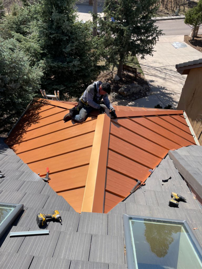 Roofer on a copper-colored metal roof, working near a wooden ridge cap. Trees and skylights in background.
