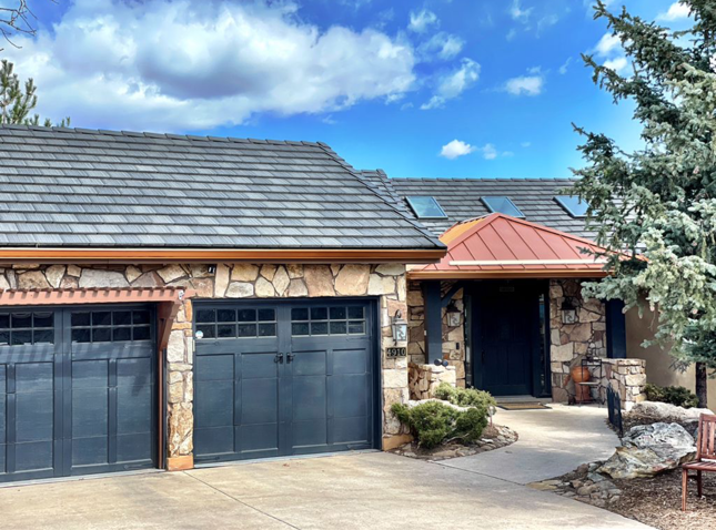 House with stone facade, blue-gray garage doors, brown roof, and copper accents on a sunny day.