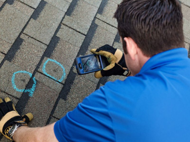 Man in blue shirt on a roof taking a photo of hail damage marked with blue chalk.