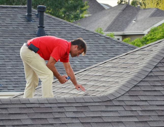 Man in red shirt inspecting a gray shingled roof. White pants, sunny setting.