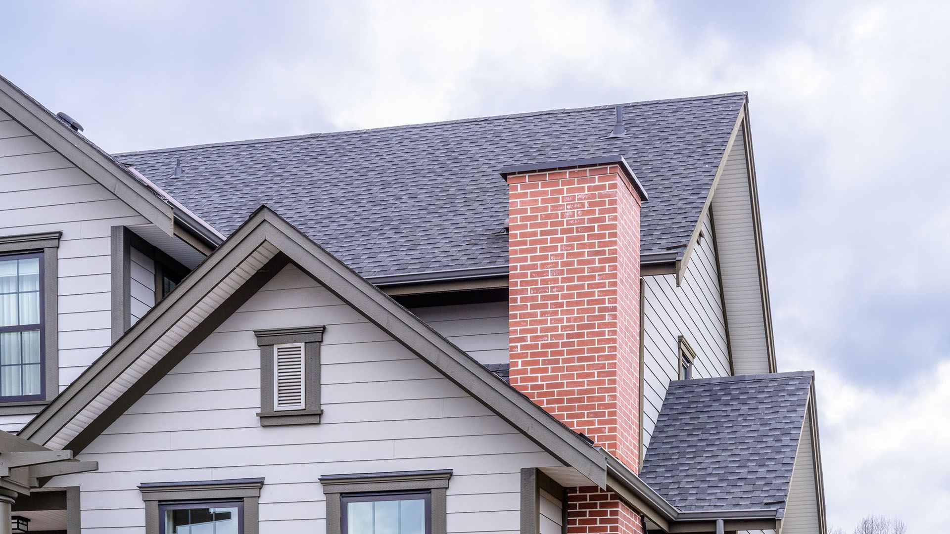 House exterior with brick chimney, gray roof, and light-colored siding against a cloudy sky.