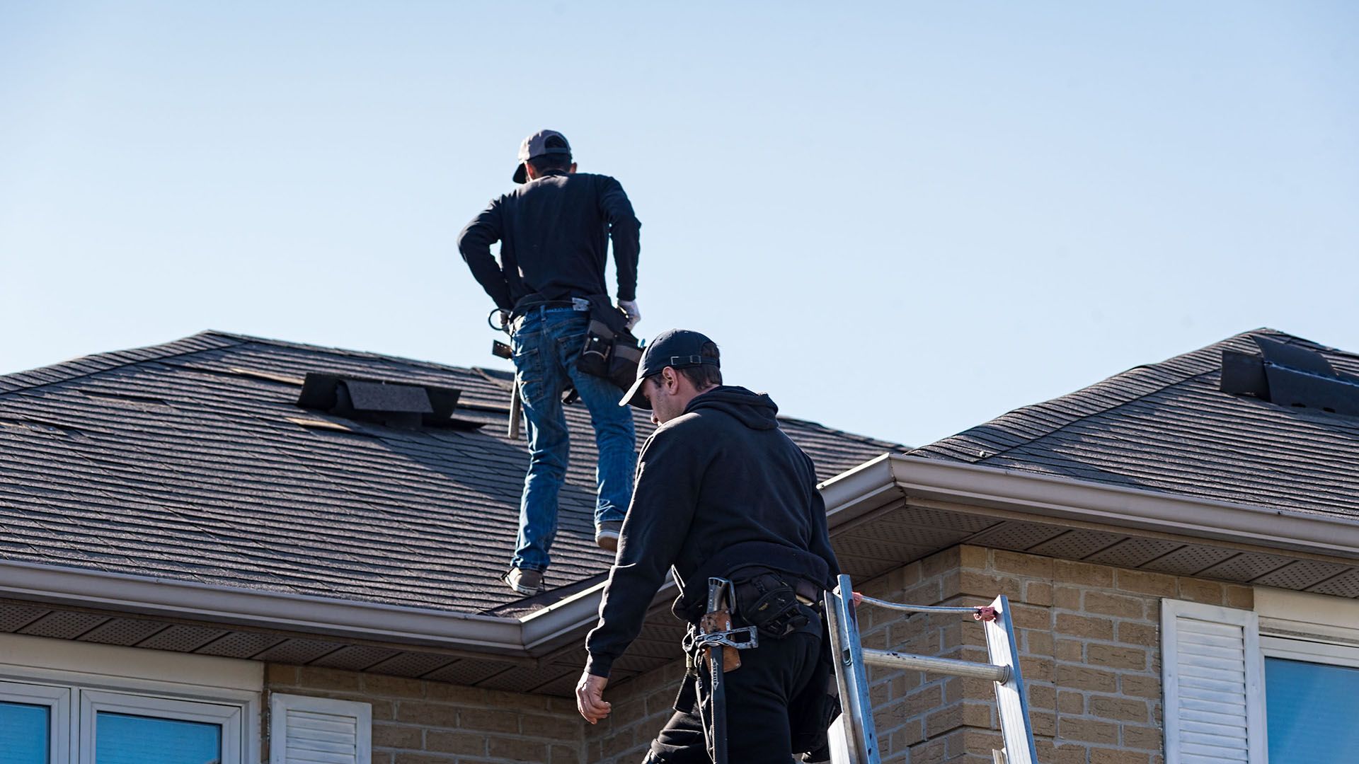 Two roofers on a rooftop, one on ladder, other on roof, working on a house on a sunny day.