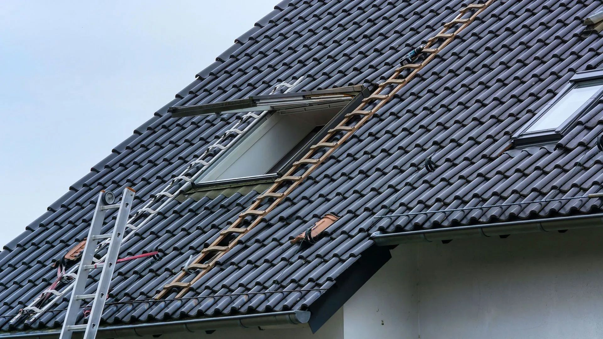 Wooden ladder on a dark tiled roof near an open skylight.