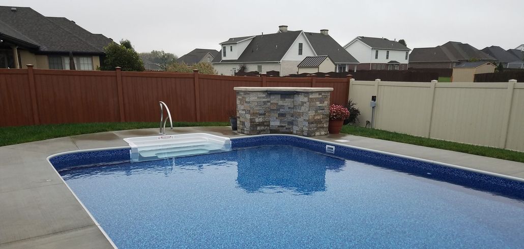 Backyard swimming pool with blue cover, stone fountain, and fenced suburban homes in the background.