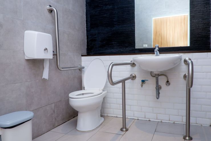 Accessible public bathroom with toilet, sink, grab bars, and paper dispenser on tiled walls.