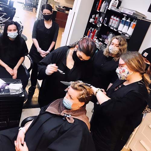 A group of women wearing face masks are standing around a woman getting her hair cut.