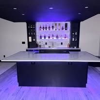 Modern bar area with white countertops, black cabinetry, purple lighting, and shelves of liquor bottles