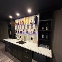 Modern bar area with illuminated liquor shelves, dark cabinets, and white countertops