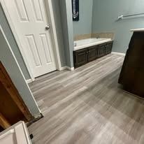 Small bathroom with gray wood-look flooring, white door, vanity, and a dark cabinet.