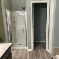 Bathroom with glass shower, vanity edge, and open doorway to a gray hallway with wood-look flooring