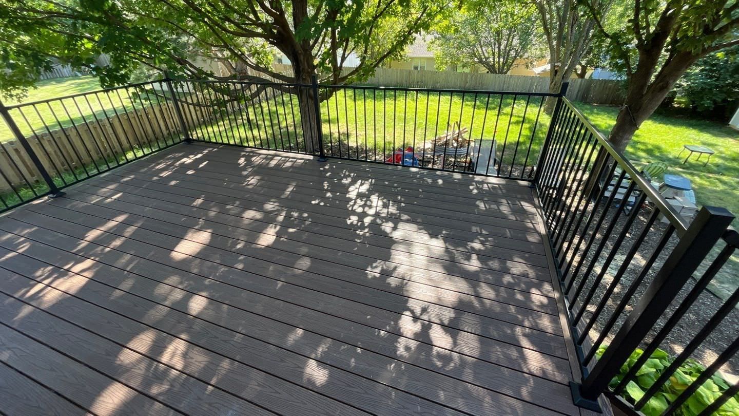 Wooden deck with black railing overlooking a grassy yard and trees, with dappled sunlight shadows.