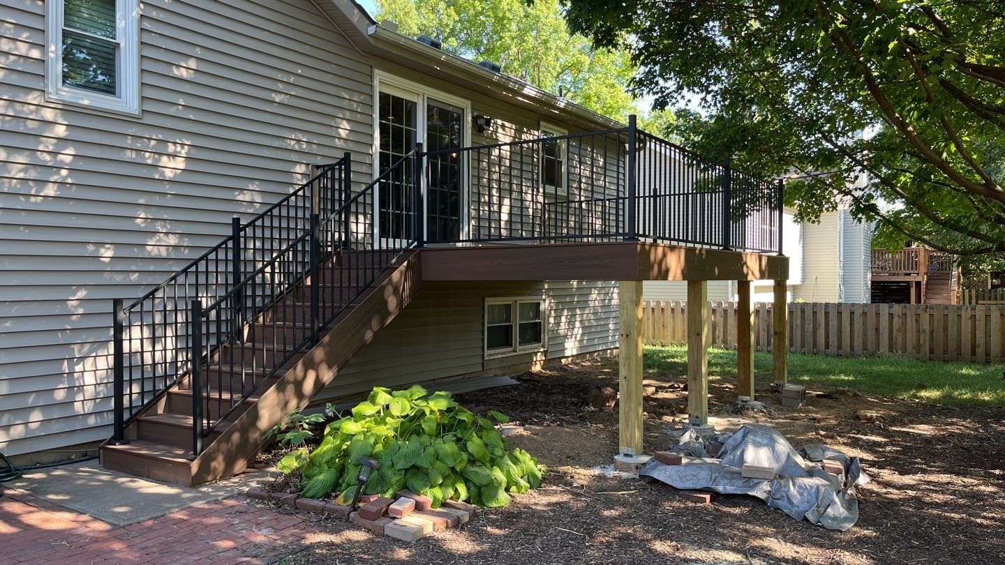 House exterior with side deck stairs, black railing, and shaded yard with rocks and trees