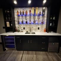 Basement wet bar with white brick backsplash, blue accent lights, shelves of bottles, and dark cabinets
