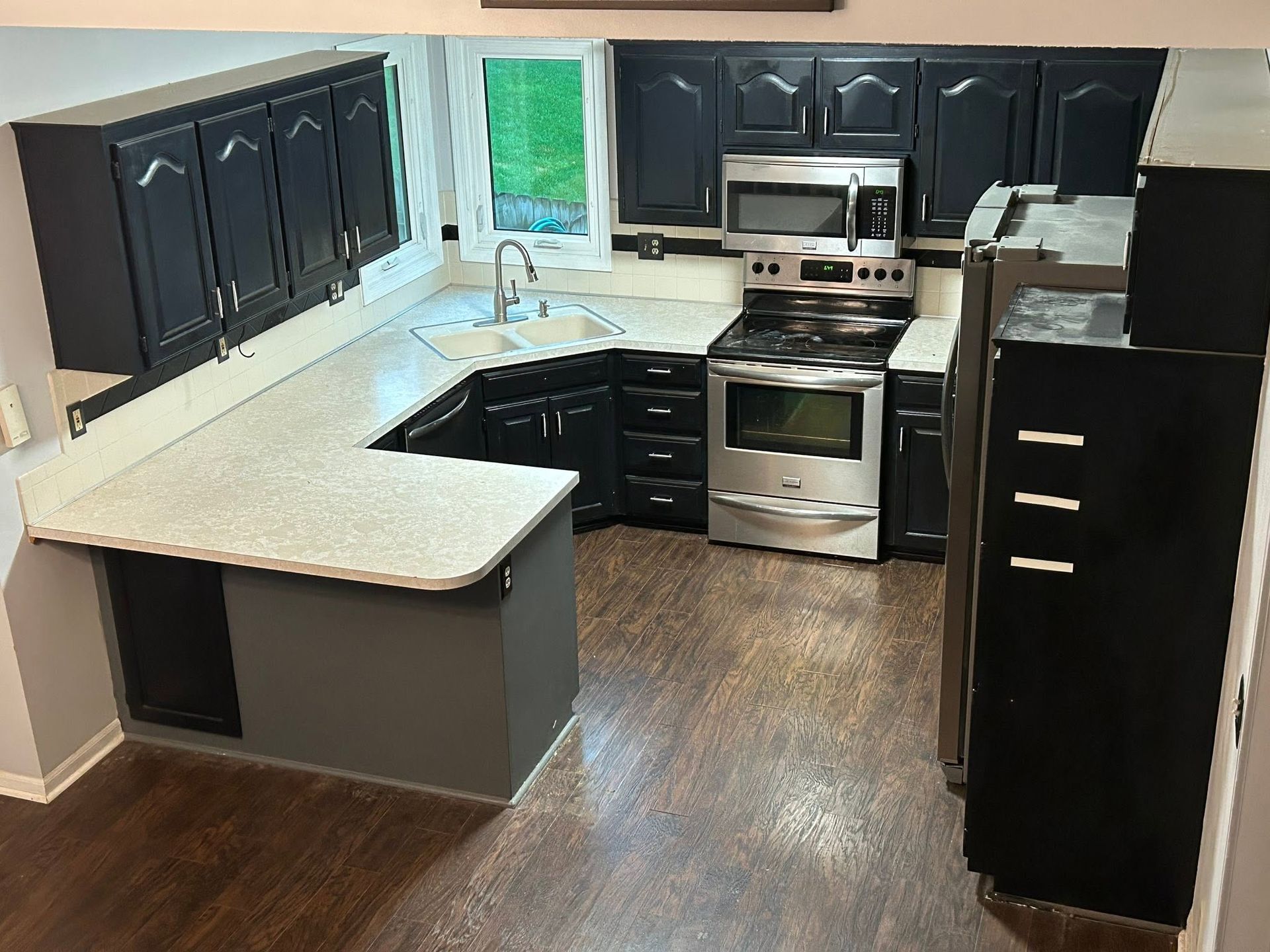 Empty modern kitchen with dark cabinets, light countertops, stainless appliances, and wood flooring.