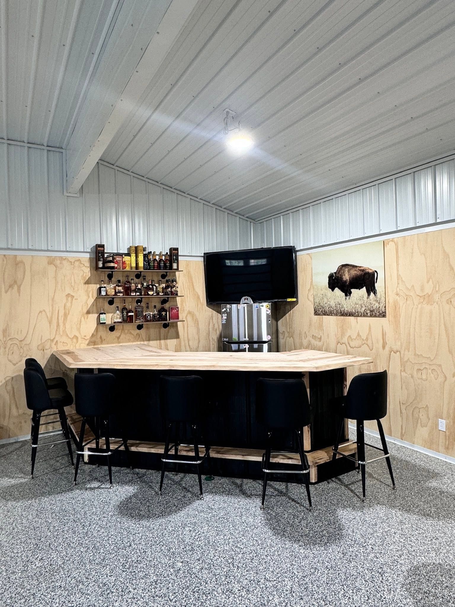 Rustic indoor bar with black counter, metal stools, wall decor, and white corrugated ceiling