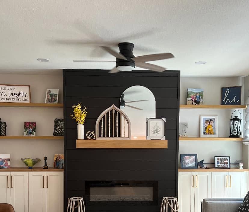 Living room fireplace with black accent wall, built-in shelves, arched mirror, and ceiling fan