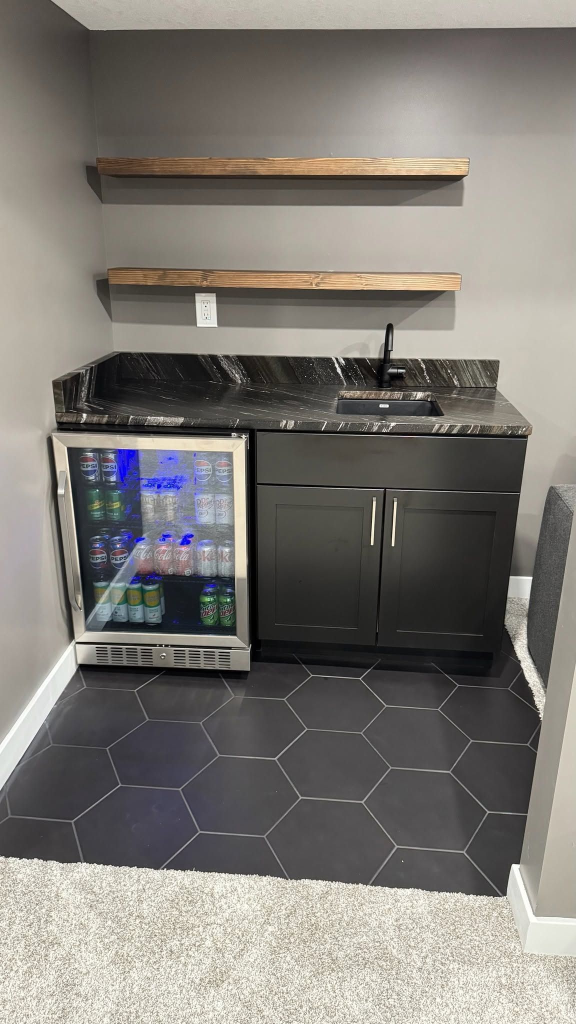 Small kitchenette with black cabinets, sink, fridge, speckled countertop, and two wooden shelves against gray walls.