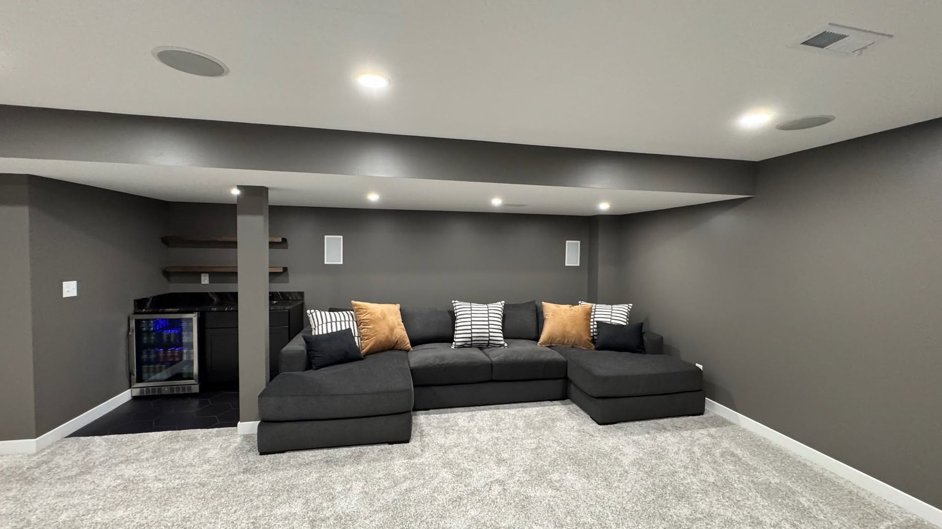 Modern basement lounge with a dark sectional sofa, gray walls, recessed lighting, and a carpeted floor.