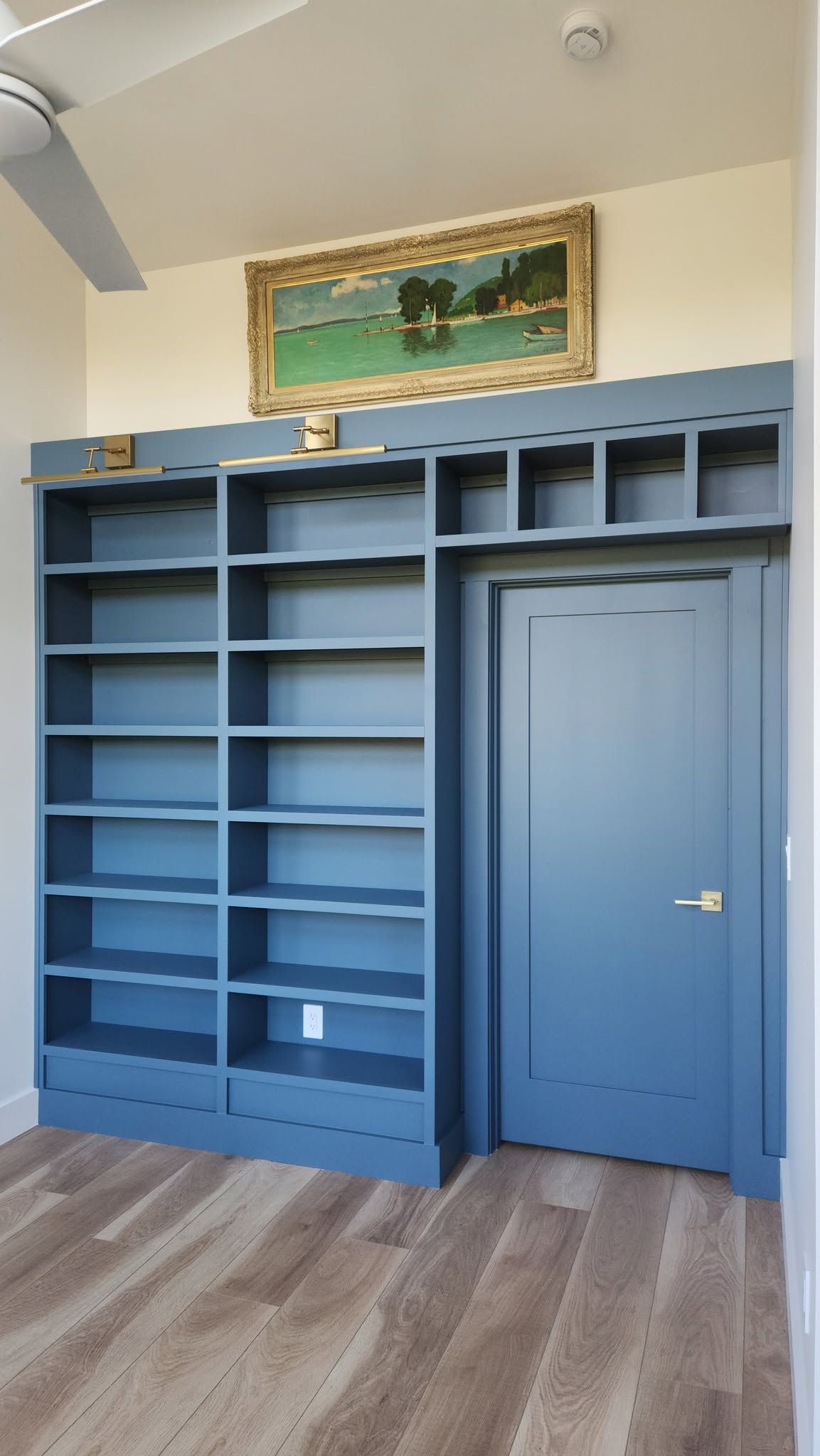 Blue built-in shelving and a closed door in a room with light wood flooring and a framed painting above.