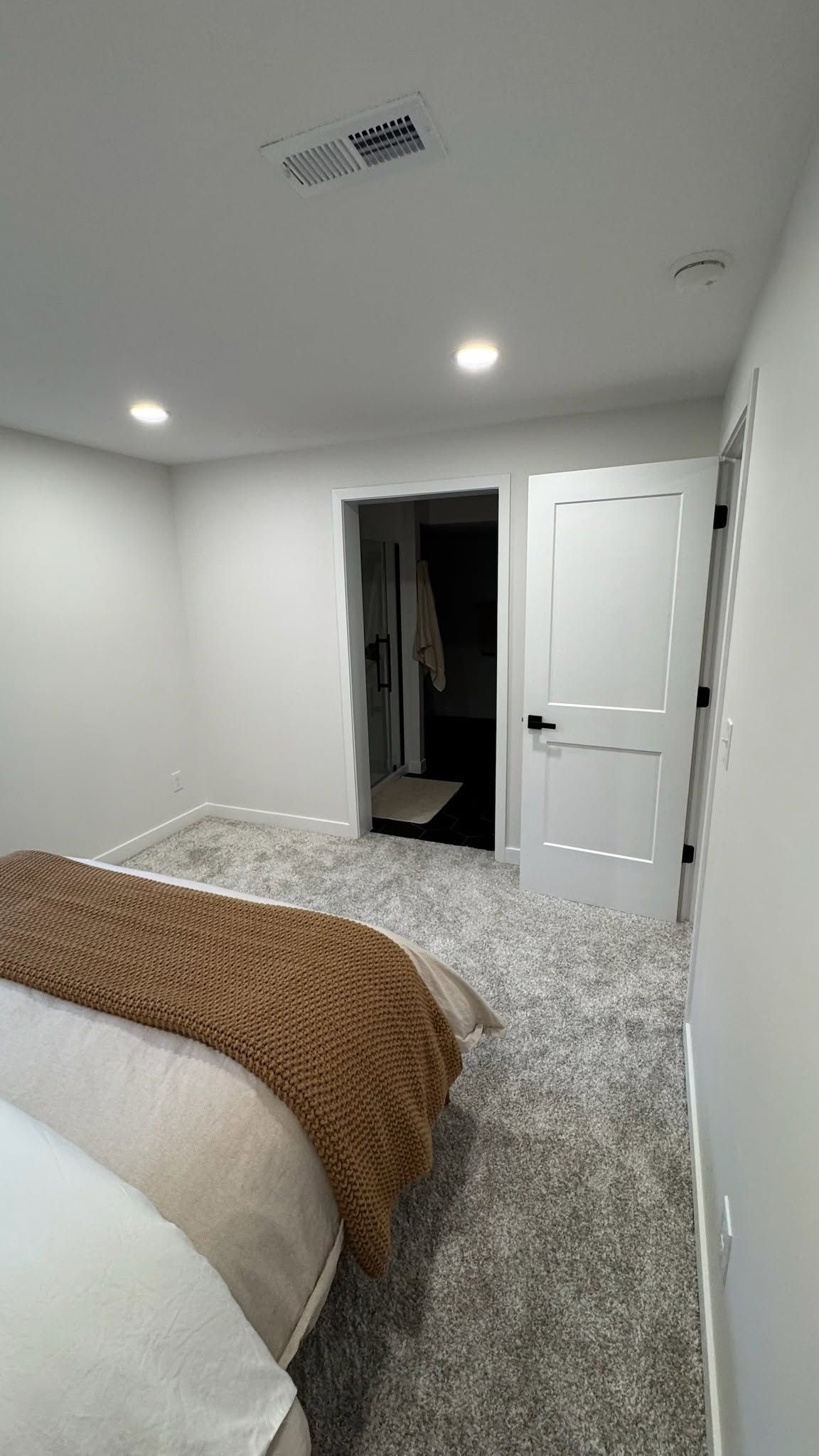 Small bedroom with gray carpet, white walls, and an open doorway; bed with patterned blanket in foreground