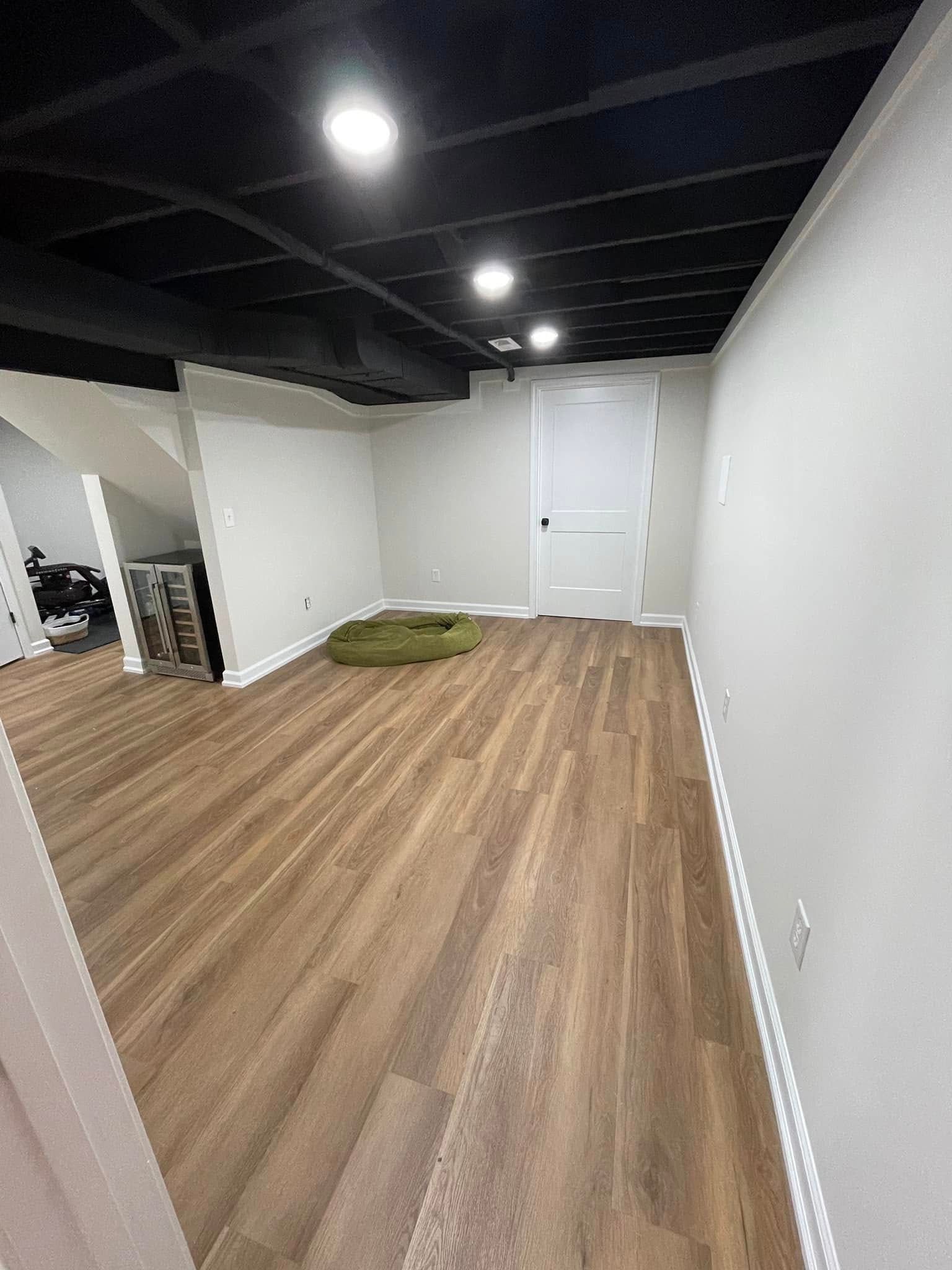 Empty white room with wood flooring, black ceiling, and a green cushion near the back wall.
