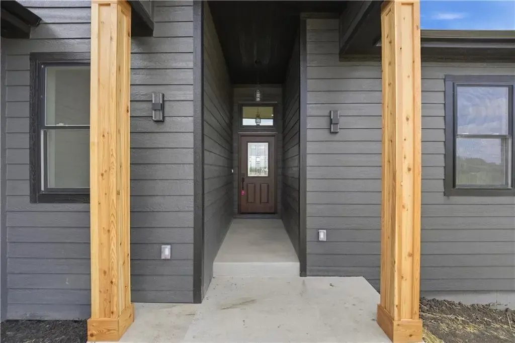 Front porch entry with gray siding, wooden posts, and a concrete walkway leading to a brown door.