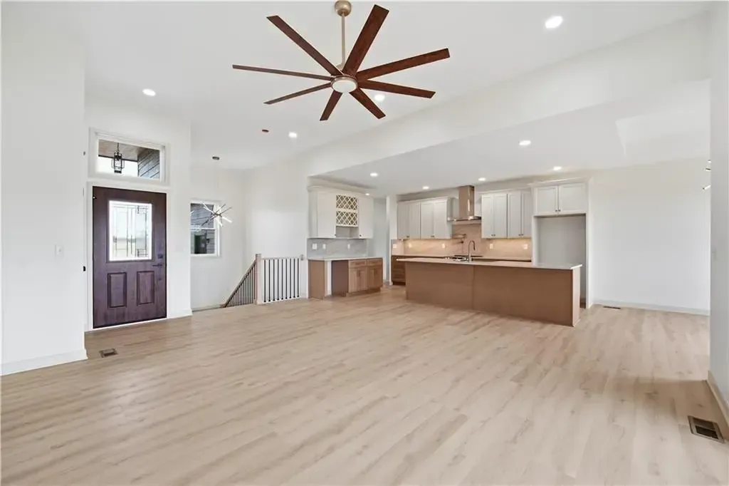 Spacious modern living room with light wood floors, white walls, ceiling fan, and open kitchen bar.