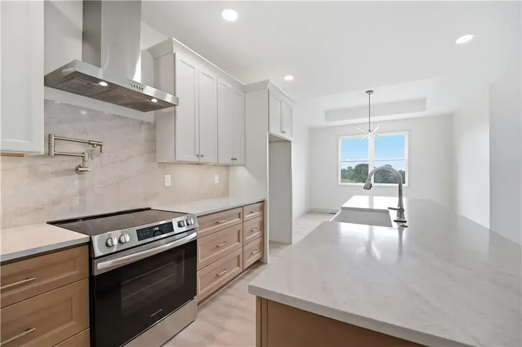 Bright modern kitchen with white cabinets, stainless stove, marble island, and dining area by large window.