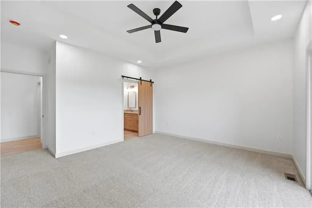 Spacious empty bedroom with white walls, carpet, ceiling fan, and open doorway to a bathroom.