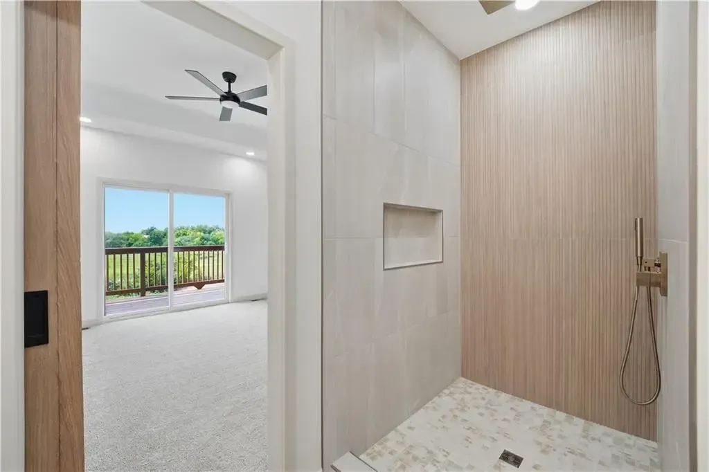 Modern bathroom with shower area, beige wood-look walls, and a view into a bright bedroom with balcony doors.