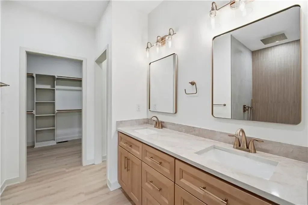 Bright bathroom vanity with double sinks, wood cabinets, mirrors, and open walk-in closet.