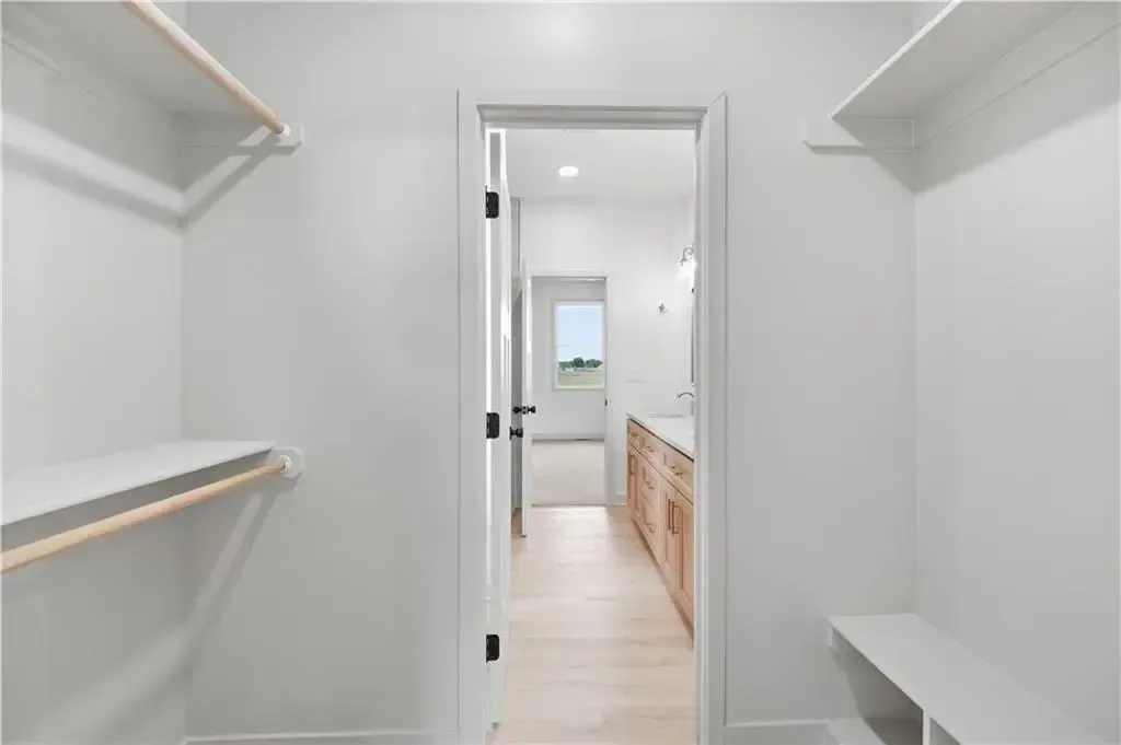 Empty walk-in closet with white shelves and a doorway to a bright bathroom beyond