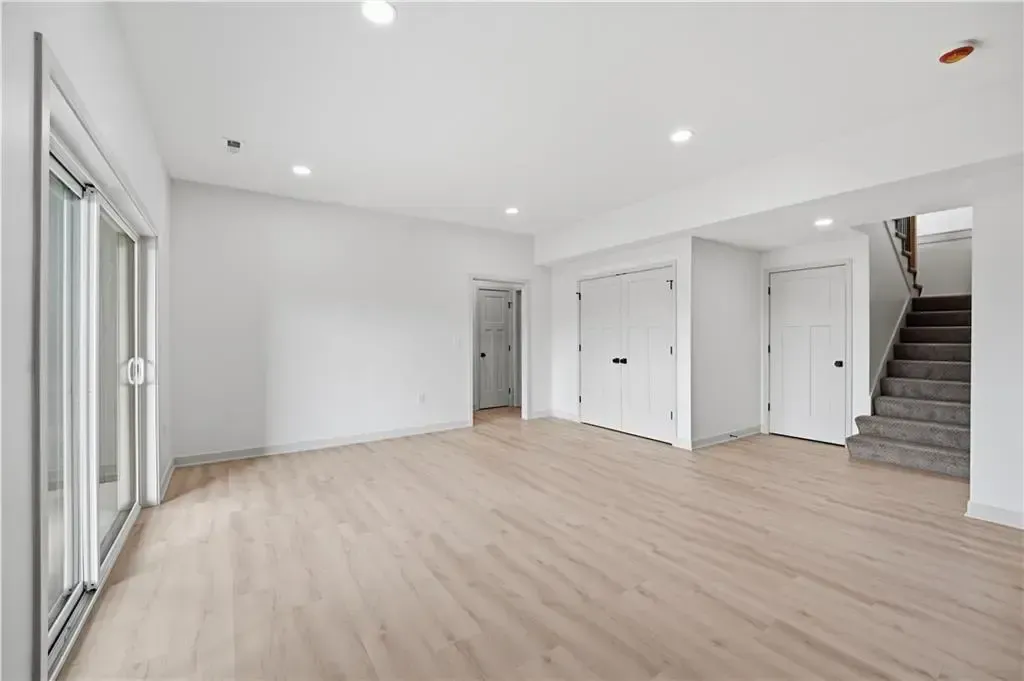 Bright empty living room with light wood floors, white walls, sliding doors, and a staircase on the right