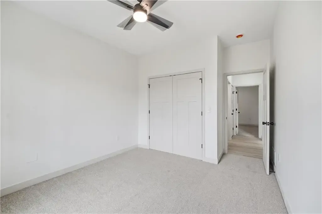 Empty white bedroom with carpet, ceiling fan, closet doors, and open doorway to a hallway.