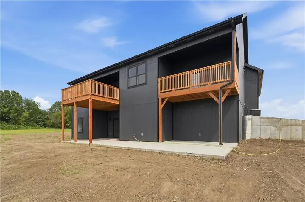 Modern two-story house with dark siding and wooden balconies on a dirt lot under a blue sky