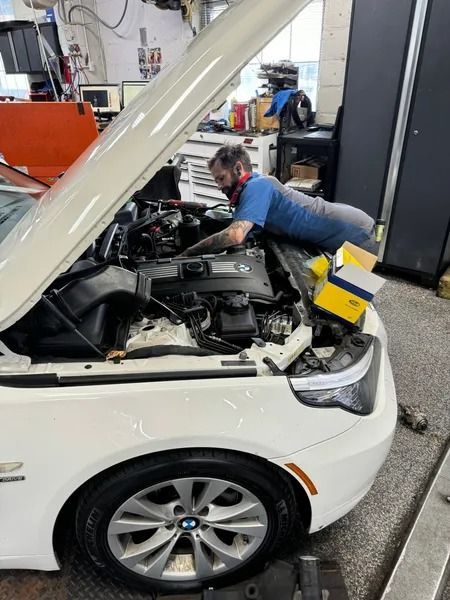 A mechanic in a blue shirt leans over the open hood of a white BMW sedan to perform repairs in an auto shop.