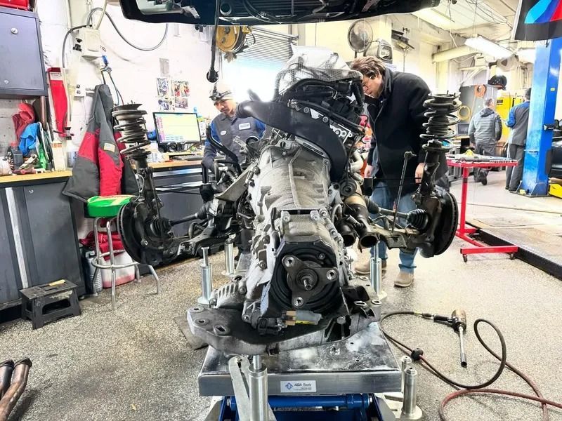 Two mechanics work on a car engine and transmission assembly mounted on a service stand in an auto repair shop.