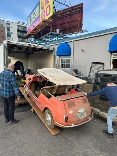 Two people push a vintage, orange, convertible microcar up a wooden ramp and into an open cargo trailer.