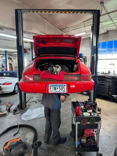 A technician works under a red Porsche on a lift in an auto repair shop with an open trunk and engine visible.