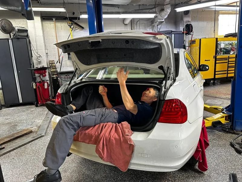 A person lying inside the open trunk of a white car in an auto repair shop, holding a tool and working on the vehicle.