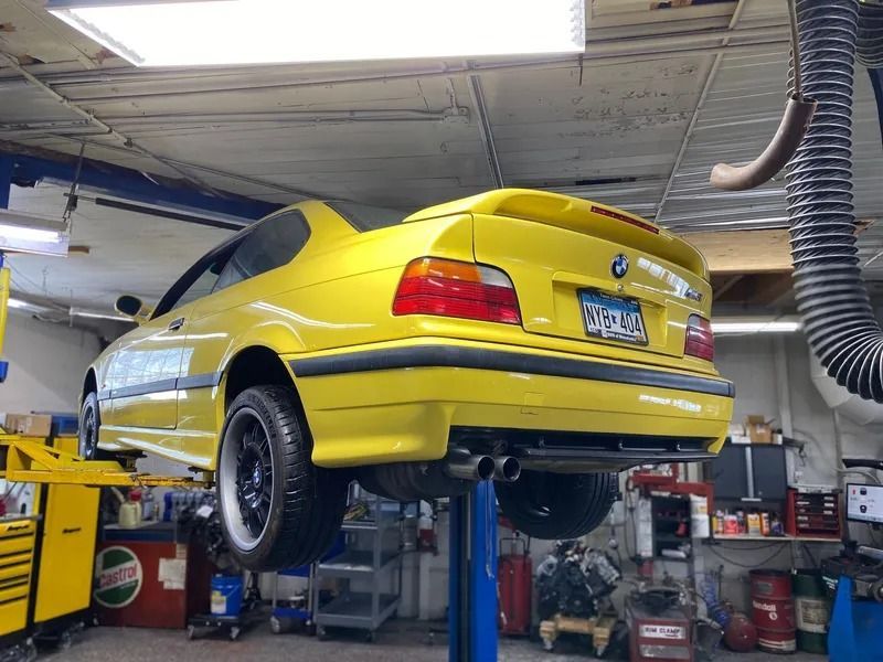 A yellow BMW E36 M3 coupe is lifted on a mechanical service rack inside an automotive repair shop.