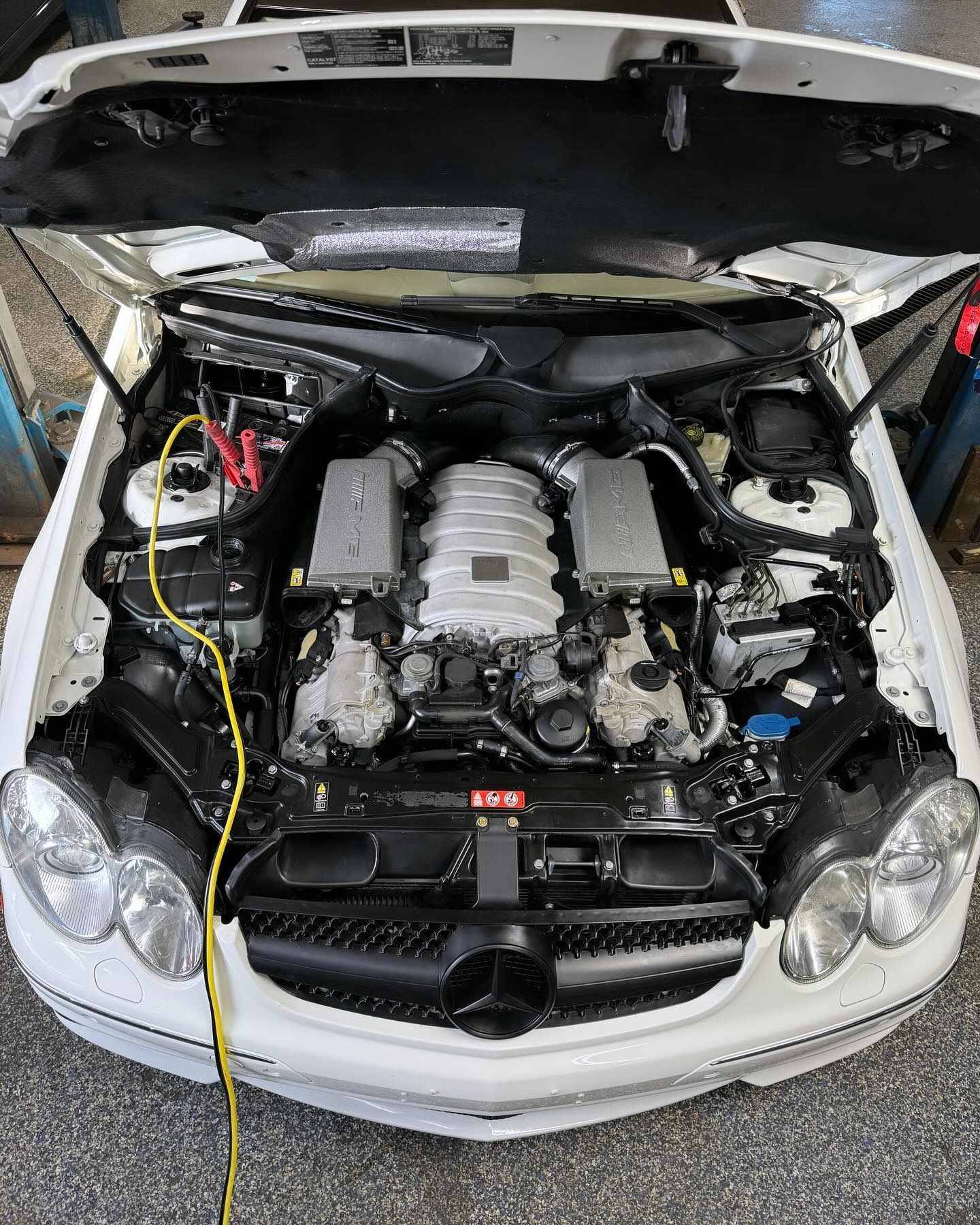 Engine bay of a white Mercedes-Benz with a yellow jump starter cable connected to the battery.