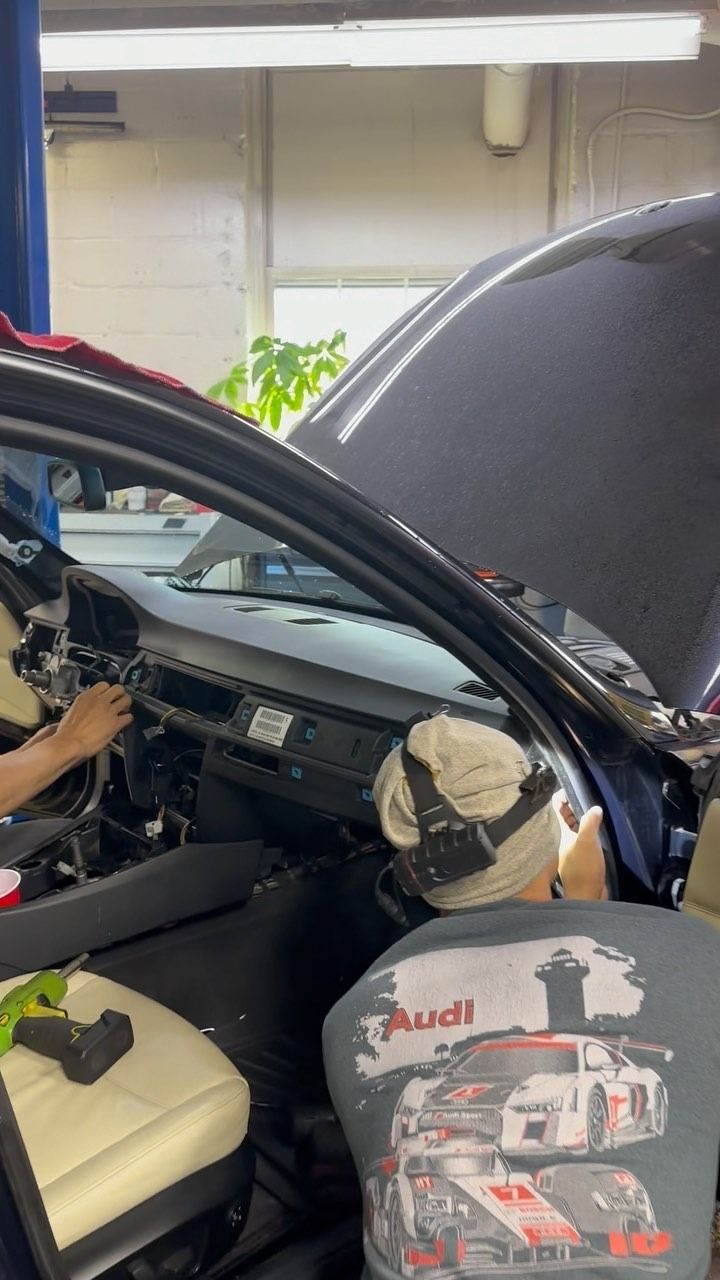 A mechanic wearing an Audi shirt repairs the interior dashboard of a car with its hood open in a garage.