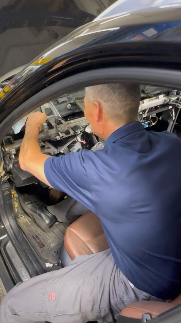 A technician in a blue shirt works on the exposed interior wiring and dashboard components of a car.