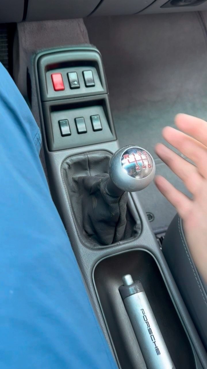A first-person view of a manual car gear shifter, center console buttons, and a Porsche-branded tire pressure gauge.