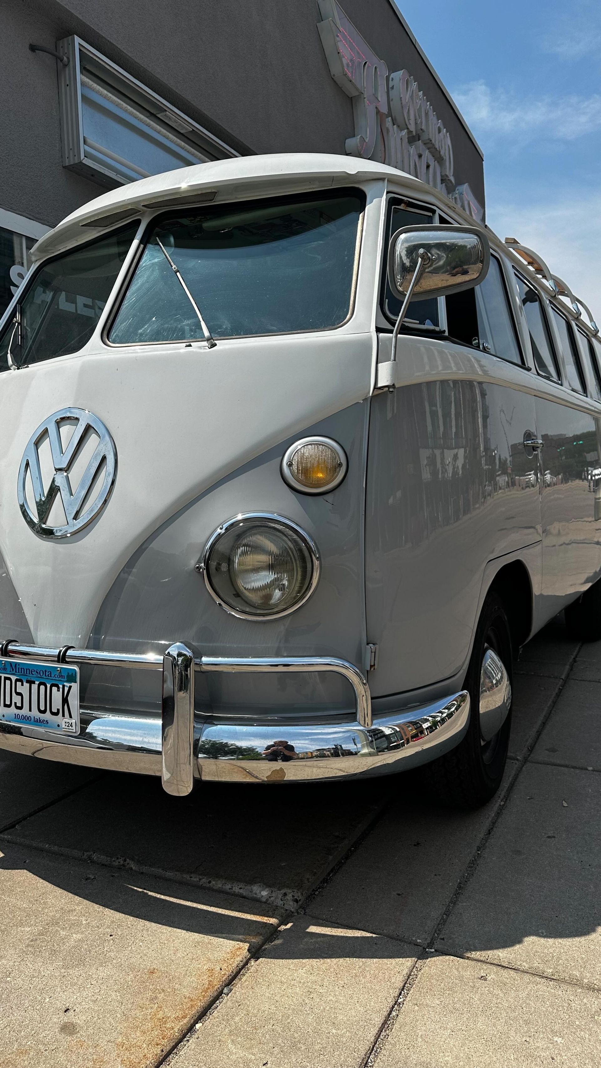 A two-tone, light gray and white vintage Volkswagen bus parked on a concrete sidewalk in front of a building.