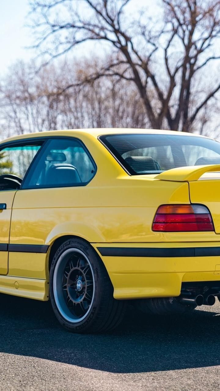 A bright yellow BMW M3 coupe parked on asphalt with bare trees in the background under a clear sky.