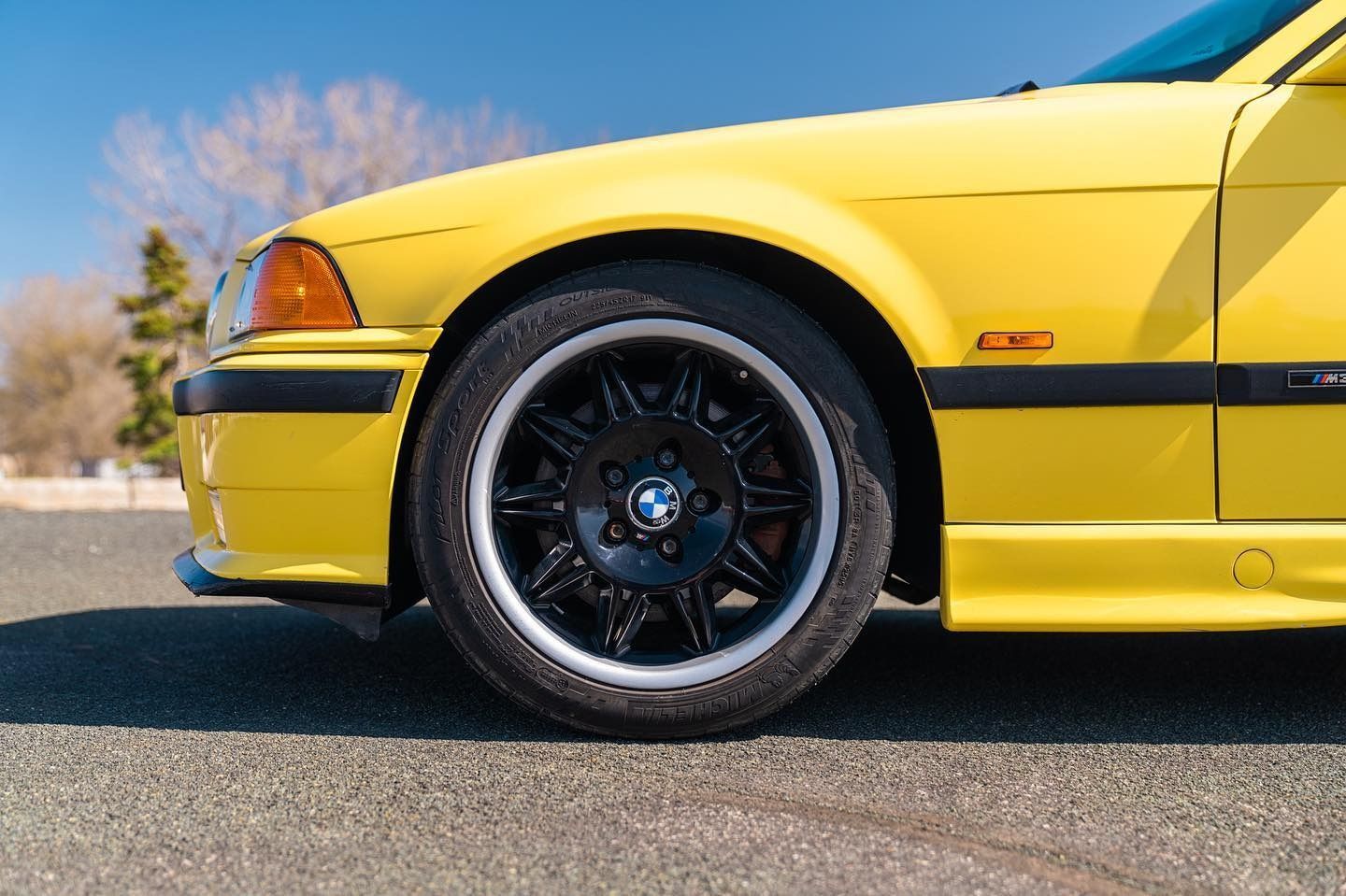 A close-up, side profile view of a yellow BMW E36 M3 parked on asphalt, highlighting the front wheel and fender.