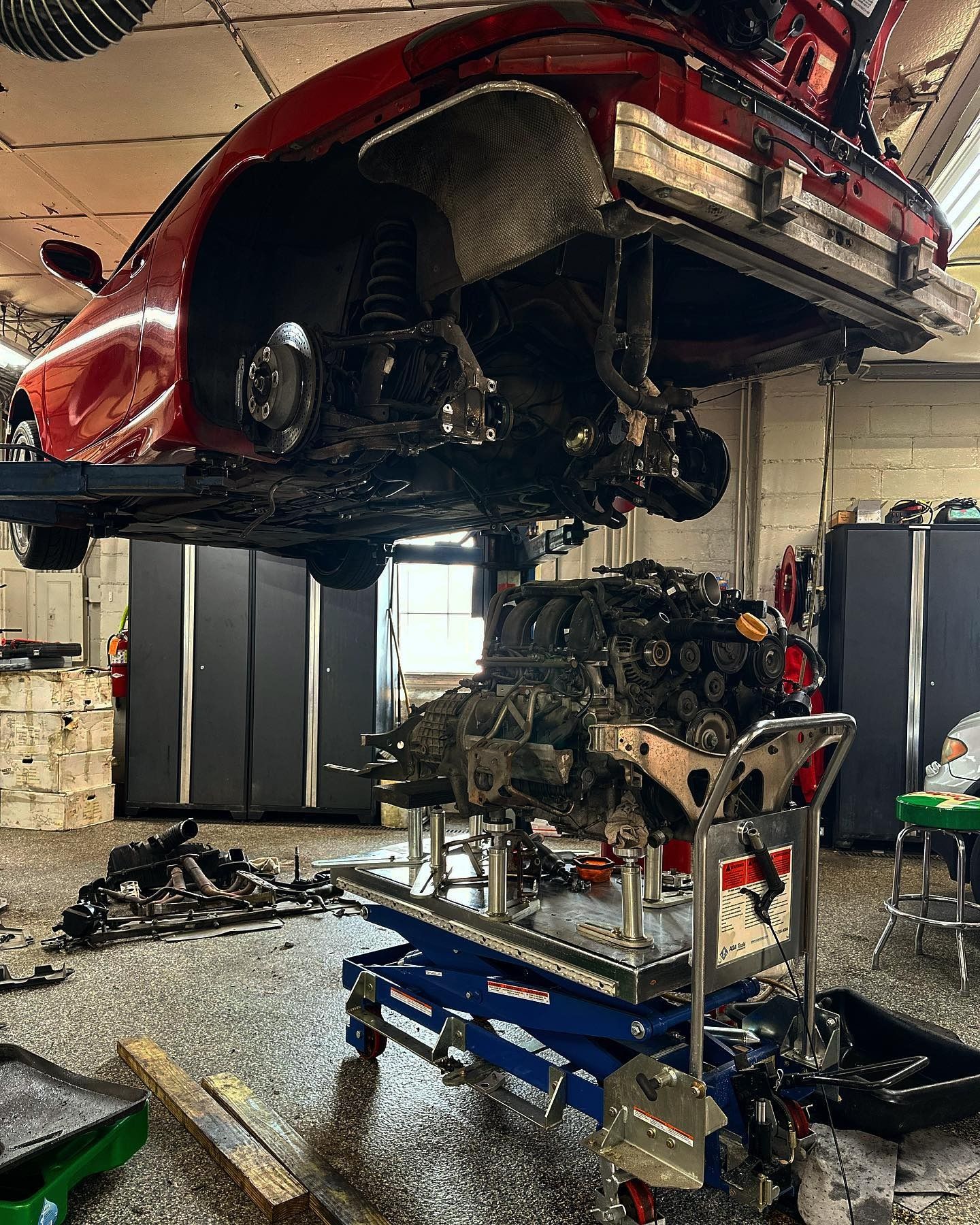 A red car body is lifted on a hoist in a garage, with its engine removed and supported on a blue rolling work table.